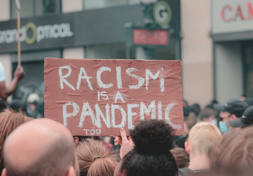 Demonstration vieler Menschen und ein Schild, auf dem steht: "Racism is a pandemic, too!"