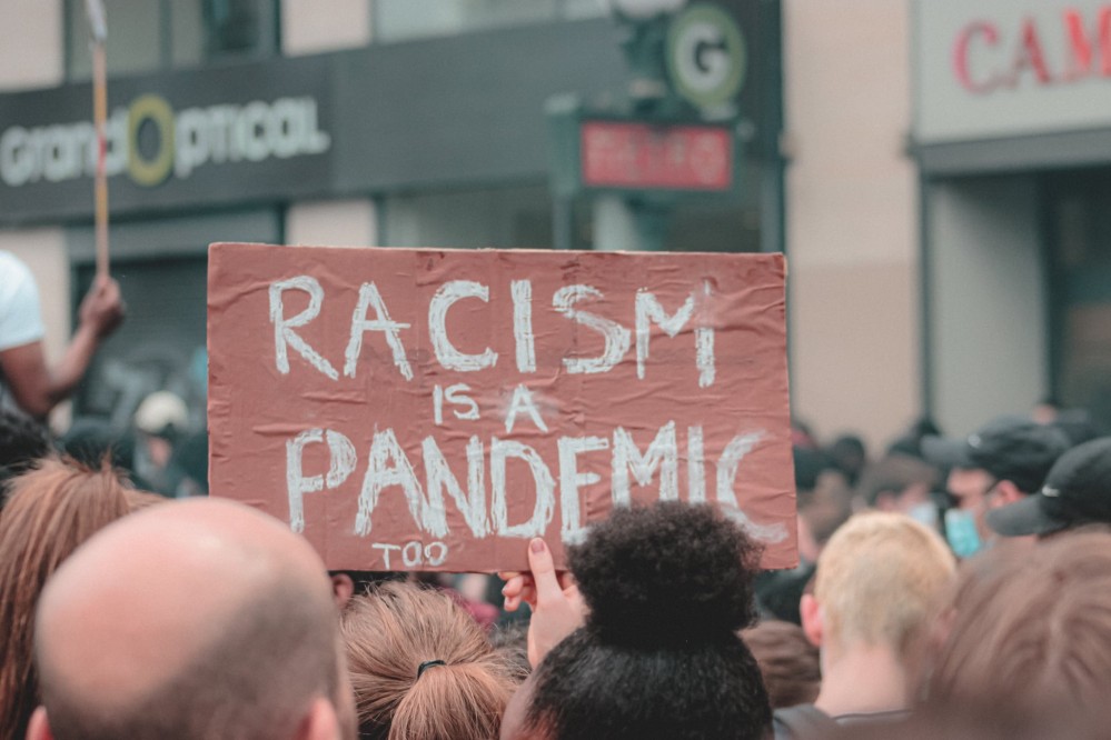 Demonstration vieler Menschen und ein Schild, auf dem steht: "Racism is a pandemic, too!"